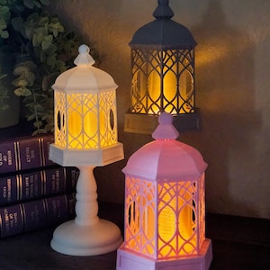 May include: Three decorative lanterns in white, gray, and pink, each with an intricate lattice design. The lanterns emit a warm, inviting glow. One lantern is on a pedestal, while the others are on a surface. The lanterns are next to a stack of books.