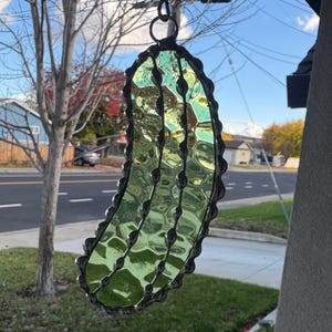 May include: A green stained glass pickle ornament with a silver metal frame. The pickle is hanging from a silver chain.