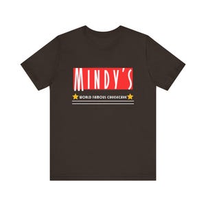 May include: A brown t-shirt with a red rectangle that says "MINDY'S" in white text. Below the rectangle is the text "WORLD FAMOUS CHEESECAKE" in white text with a yellow star on each side.