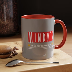May include: A gray ceramic mug with a red interior and handle. The mug features the text "MINDY'S" in a red rectangle, with "WORLD FAMOUS CHEESECAKE" below. A star is on each side of the text. A spoon and a macaron are on the wooden surface.