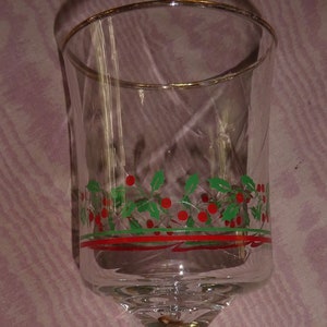 May include: A clear glass goblet with a gold rim and a decorative band of green holly leaves and red berries. The goblet has a long stem and a round base, suitable for serving beverages.