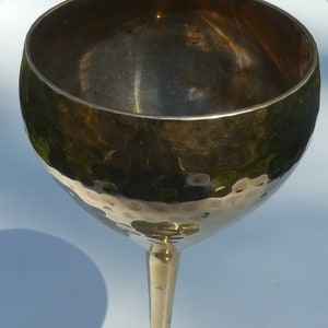 May include: A vintage champagne glass with a hammered metal finish. The glass has a wide, rounded bowl and a slender stem leading to a circular base. The metal has a warm, golden hue, reflecting light and creating a textured appearance. The glass is likely made of brass or a similar alloy.
