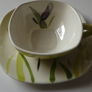May include: A white ceramic teacup and saucer set with a green and purple floral design. The cup has a square shape and a handle.