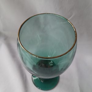 May include: A teal-colored wine glass with a gold rim. The glass has a rounded bowl and a stem with a circular base. The glass is empty and appears to be made of glass.