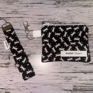 May include: A black and white dragonfly pattern decorates a wristlet keychain and a small zipped pouch. The wristlet has a metal ring and a white tag with the text "BraxDot". The pouch also has a white tag with the text "BraxDot Designs".