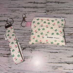May include: A set of accessories with a floral design. It includes a wristlet key fob, a small zipped pouch, and a keychain. The wristlet and pouch are cream-coloured with pink flowers and green leaves. The pouch has the text "BraxDot Designs".