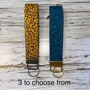 May include: Two keychains with leopard print fabric. One keychain is yellow with a gold ring, the other is blue with a silver ring. The text "3 to choose from" is at the bottom of the image.
