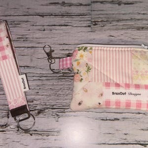 May include: A matching set featuring a pink and white gingham wristlet strap and a small zippered pouch. The pouch has a patchwork design with floral and striped patterns, and a bunny face. The brand name "BraxDot Designs" is visible.