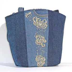 May include: A denim tote bag with gold embroidery on a central panel. The bag, with two handles, is made from various shades of blue denim. The embroidery features floral and swirling patterns.