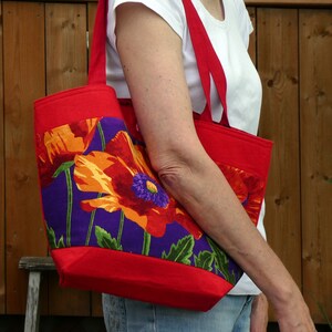 May include: A red tote bag with a floral design featuring orange, red, and purple poppies. The bag has red handles and trim. The floral pattern is set against a purple background.