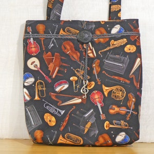 May include: A black tote bag patterned with various musical instruments in brown, gold, and red. It has a grey rope trim, a decorative button, and a silver treble clef charm. The handles are made from the same fabric. A stylish accessory.