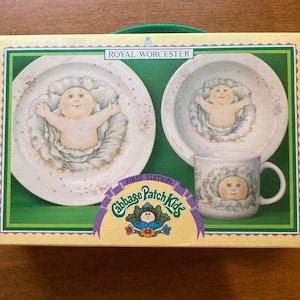 Può includere: Un set di piatti Royal Worcester Cabbage Patch Kids in scatola. Il set include un piatto, una ciotola e una tazza, ognuno con un bambino in un design a cavolo. La scatola riporta le scritte "First Edition" e "Cabbage Patch Kids".
