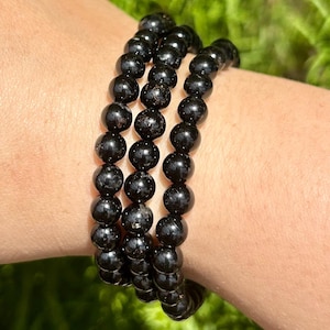 May include: Three black beaded bracelets. Each bracelet is made of round, polished black beads. The bracelets are stacked on top of each other, creating a layered effect. The beads have a glossy finish and reflect light.