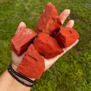 May include: A collection of four raw red jasper stones with irregular, angular shapes and matte textures. The stones exhibit a deep, solid red coloration with subtle variations in tone and natural surface fractures, held together in a palm.
