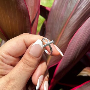 May include: A silver-colored ring with an Ankh symbol. The ring is held against a backdrop of deep red and purple leaves. The ring's design is simple and elegant, with a cross-like shape at the top.