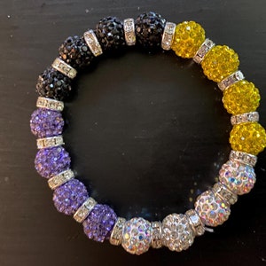 Könnte beinhalten: Ein farbenfrohes Perlenarmband mit Farbverlauf. Das Armband wechselt von Schwarz zu Lila, dann zu Gelb und schließlich zu schillernden weißen Perlen. Silberne Abstandshalter trennen die größeren Perlen.