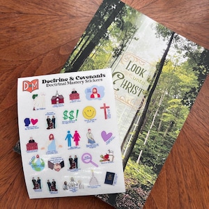 Op de afbeelding: Een vel met "Doctrine & Covenants Doctrinal Mastery Stickers" met diverse religieuze illustraties en tekst. Achter het stickervel ligt een kaart met de tekst "Look Unto Christ" en een boslandschap. De items liggen op een houten oppervlak.