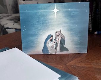 May include: A blue Christmas card featuring an illustration of the Nativity scene. The image depicts Mary, Joseph, and baby Jesus under a bright star. The card is on a wooden surface with a stack of blank cards in the foreground.
