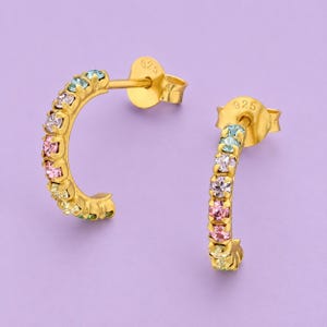 May include: A pair of gold-tone hoop earrings featuring a row of multi-colored crystals in shades of pink, yellow, and light blue. Each earring has a post back closure stamped with 925, indicating sterling silver construction with a gold finish.