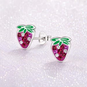 May include: A pair of silver stud earrings shaped like strawberries. Each strawberry has a red and pink crystal-like design with green leaves at the top. The earrings are on a sparkly, light-colored background.