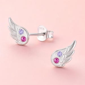 Puede incluir: Un par de pendientes de plata de ley con forma de ala, cada uno con una pequeña piedra preciosa redonda de color morado y otra de color rosa. La parte posterior del poste del pendiente tiene grabado el número 925, indicando el contenido de plata.
