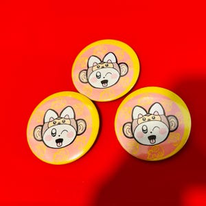 May include: Three yellow and pink round buttons with a cartoon monkey wearing a hat. The monkey has a pink face with a big smile and a pink tongue sticking out. The monkey is wearing a brown hat with a white face and a small smile.
