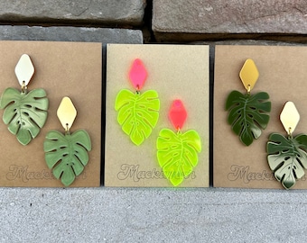 May include: Three pairs of earrings featuring a green monstera leaf design. The earrings are displayed on brown card stock with the word "Mackenzie" written on each card. The earrings are made of acrylic and have a gold-tone metal finding.