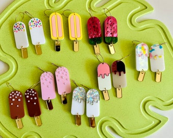 Puede incluir: Un conjunto de 10 pares de pendientes coloridos con forma de paleta de helado. Los pendientes están hechos de arcilla polimérica y tienen una variedad de diseños, que incluyen chispas, sandía y purpurina. Los pendientes están todos en aros de oro.