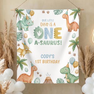 May include: A white banner with a dinosaur theme, featuring illustrations of various dinosaurs, palm trees, and balloons. Text on the banner reads "Our Little Dino is a One A-Saurus!" and "Cody's 1st Birthday."