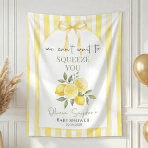 May include: A baby shower banner with a lemon theme. The banner has yellow and white stripes and features the text "we can't wait to SQUEEZE YOU" and the name "Olivia Snyder's BABY SHOWER 10.10.2026". Gold and white balloons are in the background.