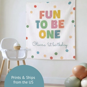 K&ouml;nnte beinhalten: Ein wei&szlig;er Wandbehang aus Stoff mit dem farbigen Text FUN TO BE ONE und dem in Schreibschrift gehaltenen Text Oliver's 1st birthday. Das Design zeigt verstreute Punkte in Rot, Orange, Gelb, Gr&uuml;n und Blau auf der strukturierten Oberfl&auml;che.