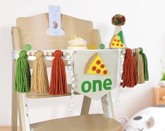 May include: A high chair decorated for a first birthday party with a pizza-themed banner, a pizza hat, and green, tan, and orange tassels.