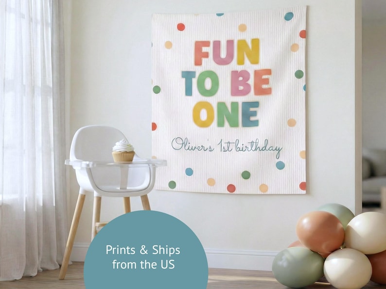 K&ouml;nnte beinhalten: Ein wei&szlig;er Wandbehang aus Stoff mit dem farbigen Text FUN TO BE ONE und dem in Schreibschrift gehaltenen Text Oliver's 1st birthday. Das Design zeigt verstreute Punkte in Rot, Orange, Gelb, Gr&uuml;n und Blau auf der strukturierten Oberfl&auml;che.