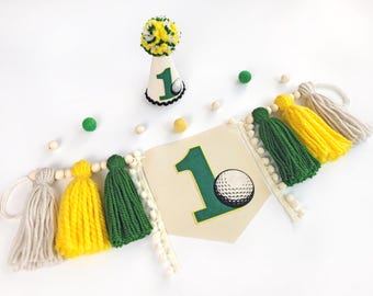 May include: A golf-themed first birthday party banner with a green number one and a golf ball. The banner is decorated with white, yellow, and green yarn tassels.