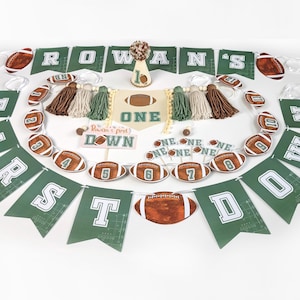 Può includere: Un set di decorazioni per feste a tema football. Include uno striscione con il nome "Rowan's", bandierine, decorazioni a forma di pallone da football e un cake topper. La combinazione di colori è verde, marrone e bianco. Lo striscione recita "Rowan's first down".