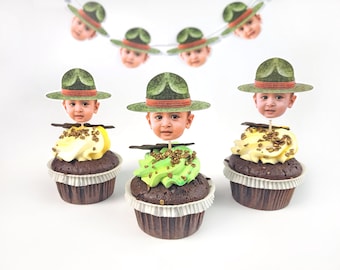 Puede incluir: Tres cupcakes de chocolate con envoltorios de papel blanco, cubiertos con glaseado verde y amarillo, y la cara de un bebé con un sombrero de guardabosques. Una pancarta con imágenes similares cuelga arriba.