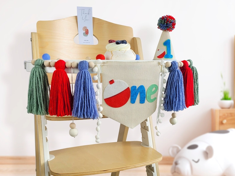 May include: A white high chair with a felt banner that says "one" with a red and white fishing bobber. The banner is decorated with green, red, and blue tassels and white pom-poms. A white and blue party hat with a red number one is on the chair.