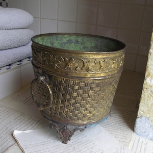May include: A round brass bucket featuring a woven texture on the exterior, a decorative band with embossed leaf patterns near the rim, two side ring handles, and three ornate feet. The interior of the bucket has a weathered green patina.