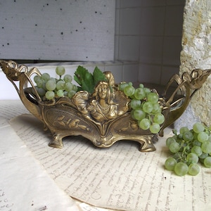 May include: An elongated brass centrepiece bowl featuring a central relief sculpture of a female figure with flowing hair, surrounded by intricate floral motifs and scrolling edges, filled with a cluster of fresh green grapes and leaves.