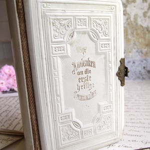 May include: A vintage white book with embossed floral and geometric designs. The cover features the German text "Andenken an die erste heilige Communion" in elegant script. A bronze clasp secures the book.