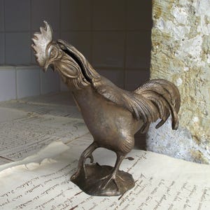 Könnte beinhalten: Eine bronzefarbene Hahnfigur mit einem Münzschlitz auf dem Rücken. Die Skulptur hat detaillierte Federn und einen markanten Kamm. Sie steht auf einem blattförmigen Sockel, platziert auf Vintage-Papier mit handschriftlichem Text.