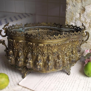 May include: An oval brass planter featuring a decorative scalloped rim and ornate handles on each side. The exterior is embossed with a repeating pattern of figures in relief, and the vessel is supported by four small, detailed metal feet.