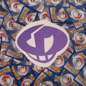 Może przedstawiać: Naklejka w kolorze fioletowym i białym z ozdobnym logo. Naklejka znajduje się na stosie niebieskich i białych kart kolekcjonerskich Pokemon.