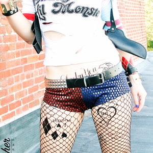 May include: A woman dressed as Harley Quinn, wearing a red and blue costume with a black leather harness and a fake gun. She has a tattoo on her stomach that says "Daddy's Lil Monster" and another tattoo on her leg that says "Joker" with a diamond and heart symbol.