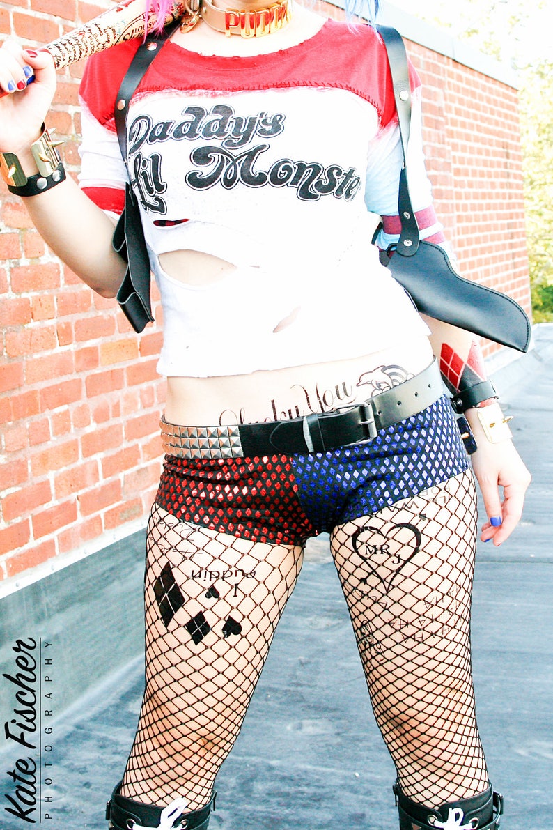 May include: A woman dressed as Harley Quinn, wearing a red and blue costume with a black leather harness and a fake gun. She has a tattoo on her stomach that says "Daddy's Lil Monster" and another tattoo on her leg that says "Joker" with a diamond and heart symbol.