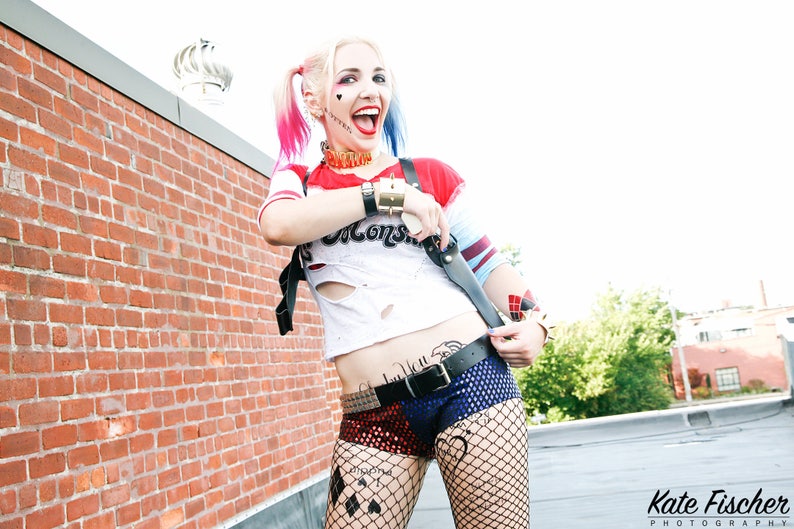 May include: A woman dressed as Harley Quinn, a comic book character, is standing in front of a brick wall. She is wearing a red and blue costume with a white shirt that has a hole in it. She has a black belt with a silver buckle and black fishnet stockings. She is holding a black and silver weapon in her right hand. She has red and blue hair and is smiling.