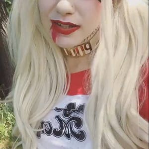 May include: A woman dressed as Harley Quinn with blonde hair, red and black clothing, and a white shirt with the text "Puddin'" on it. She is wearing makeup and has a red and black bandana in her hair.