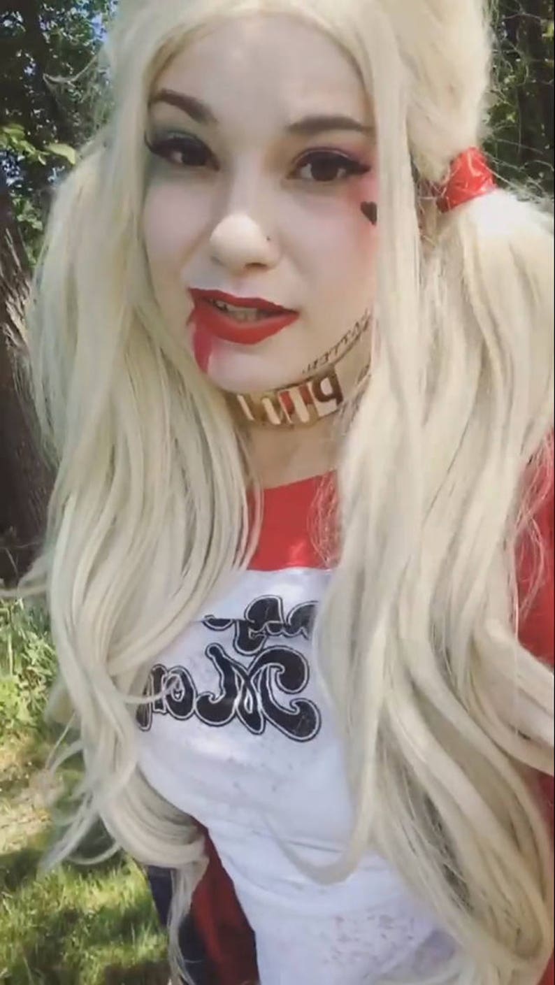 May include: A woman dressed as Harley Quinn with blonde hair, red and black clothing, and a white shirt with the text "Puddin'" on it. She is wearing makeup and has a red and black bandana in her hair.
