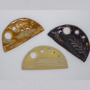 May include: Three ceramic herb markers in shades of brown, black, and gold. Each marker has a semi-circular shape with holes and embossed botanical designs. The word "HERBS" is inscribed on each marker.