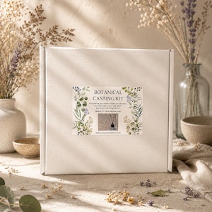 May include: A white box featuring a "BOTANICAL CASTING KIT" label with botanical illustrations. The kit contains everything needed to create and display a cast. Dried flowers and vases are in the background.